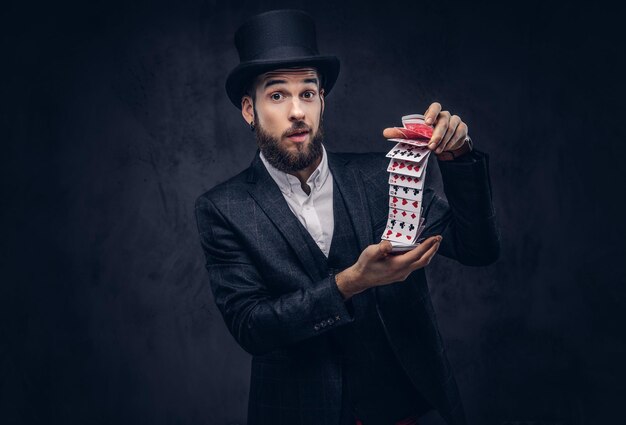 Бородатый фокусник в черном костюме и цилиндре показывает трюк с игральными картами на темном фоне.