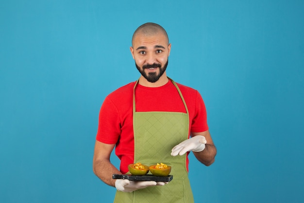 Bearded male chef in apron and gloves holding a dark wooden board with pastry . 