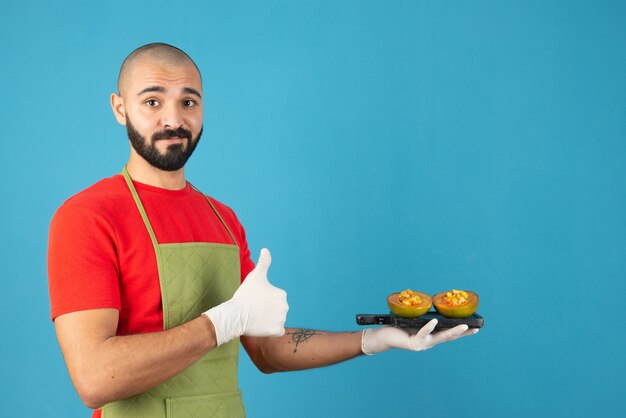 Bearded male chef in apron and gloves holding a dark wooden board with pastry . 