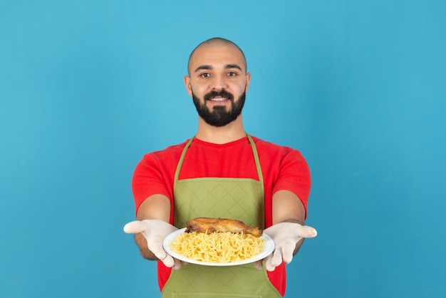 Bearded male chef in apron and gloves holding a white plate of delicious pasta with chicken meat . 