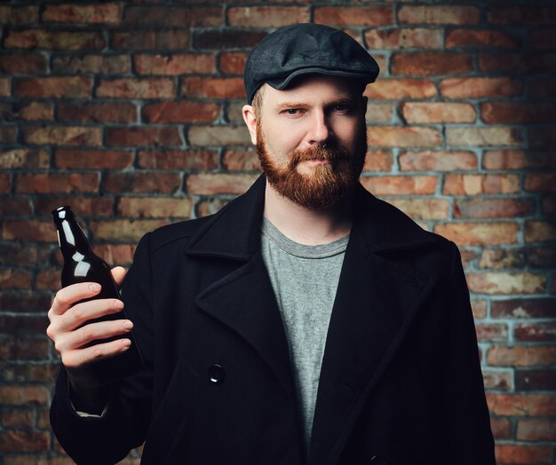 黒のツイードのフラットキャップとウールのジャケットに身を包んだひげを生やした男性は、レンガの壁にビール瓶を保持します。