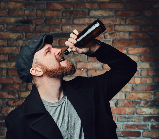 Free photo bearded male dressed in a black tweed flat cap and wool jacket holds beer bottle over the wall of a brick.