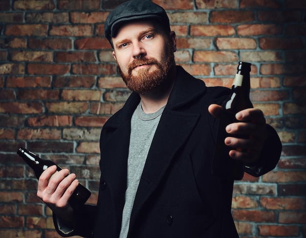 Бородатый мужчина, одетый в черную твидовую кепку и шерстяную куртку, держит бутылку пива над кирпичной стеной.