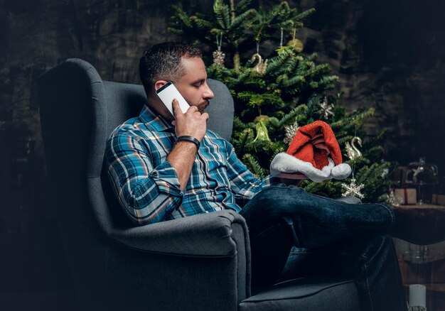 Free photo bearded male dressed in a blue plaid shirt sits on a chair and talks by smartphone over christmas decorated background.