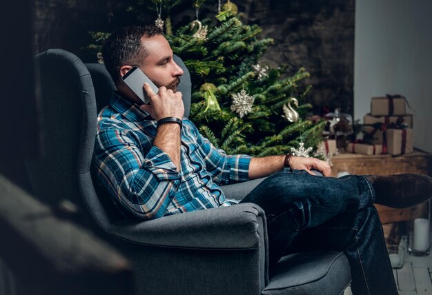 Бородатый мужчина, одетый в синюю клетчатую рубашку, сидит на стуле и разговаривает по смартфону на рождественском фоне.