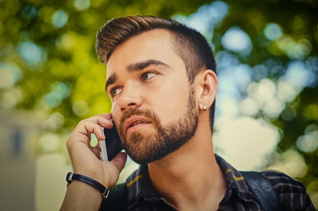 Bearded male dressed in a fleece shirt talks by a smart phone in a park.