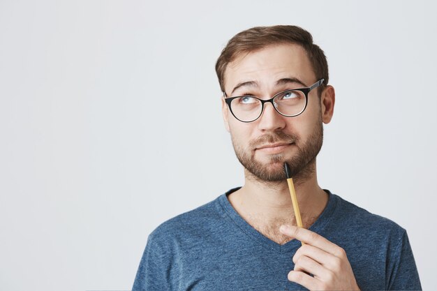Bearded male employee in glasses, holding pencil, look away pondering