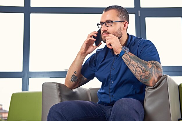 彼の腕に入れ墨をした眼鏡のひげを生やした男性は、椅子に座ってスマートフォンを使用しています。