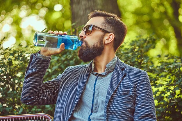 A bearded male man drinking cool water outdoors, sitting on a bench in a city park.