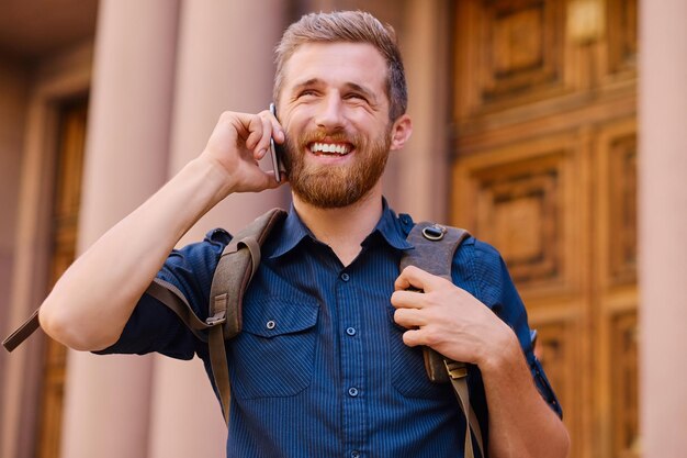 古いヨーロッパの町でスマートフォンで話すバックパックを持つひげを生やした男性。