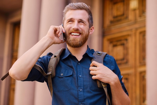 Бородатый мужчина с рюкзаком разговаривает по смартфону в старом европейском городе.