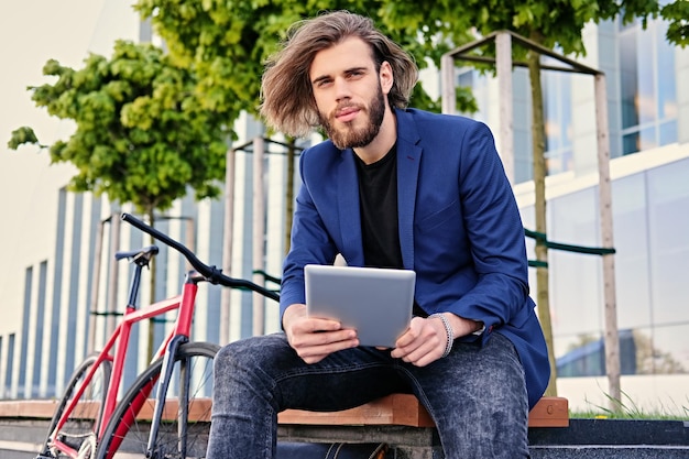 bearded male with long blond hair holds tablet PC with red single speed bicycle in a park on background.