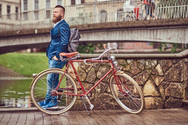 Бородатый мужчина со стильной стрижкой, одетый в повседневную одежду с рюкзаком, стоит с ретро-велосипедом у реки в городском парке.