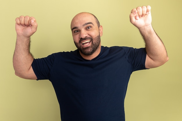 Bearded man in black t-shirt crazy happy and excited screaming clenching fists rejoicing his success standing over green wall