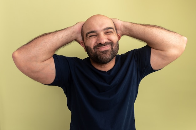 Foto gratuita uomo barbuto in maglietta nera che sembra infastidito e irritato con le mani dietro la testa in piedi sopra il muro verde