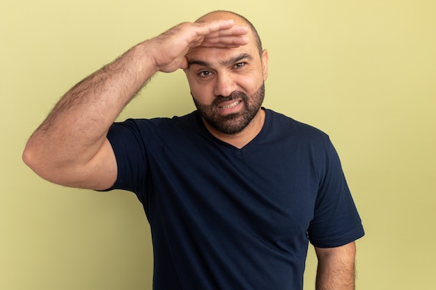 Bearded man in black t-shirt looking far away with hand over head smiling confused standing over green wall