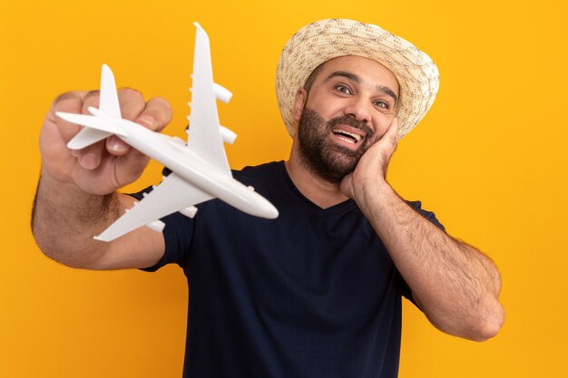 Bearded man in black t-shirt and summer hat holding toy airplane  amazed and happy smiling cheerfully standing over orange wall