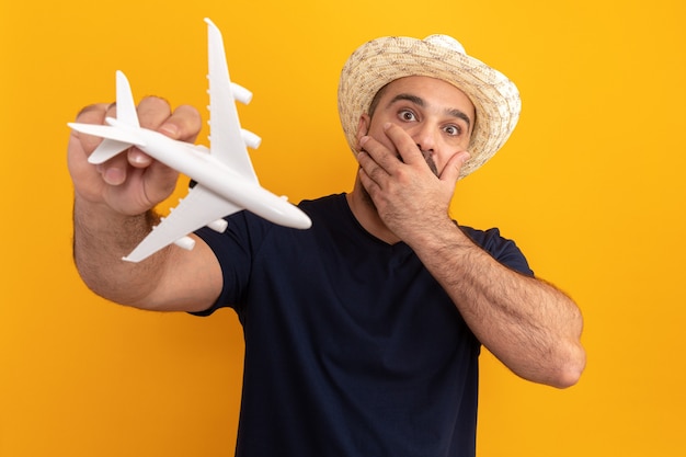 Bearded man in black t-shirt and summer hat holding toy airplane  amazed and worried covering mouth with hand standing over orange wall