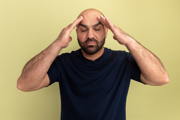 Бородатый мужчина в черной футболке, касаясь головы, выглядит нездоровым и страдает от головной боли, стоя у зеленой стены