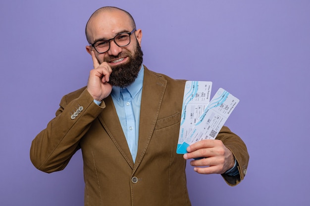 Bearded man in brown suit wearing glasses holding air tickets looking at camera smiling cheerfully happy and positive standing over purple background