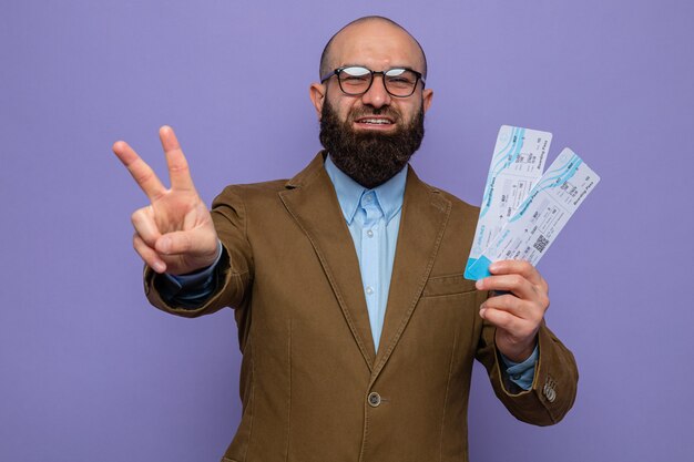 Bearded man in brown suit wearing glasses holding air tickets looking smiling cheerfully showing v-sign