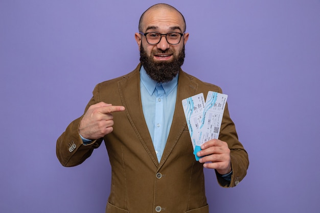 Bearded man in brown suit wearing glasses holding air tickets pointing with index finger at them smiling cheerfully happy and positive
