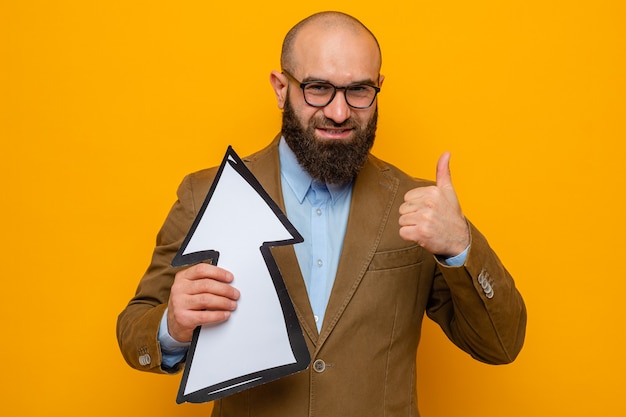 Bearded man in brown suit wearing glasses holding arrow looking at camera smiling cheerfully showing thumbs up standing over orange background