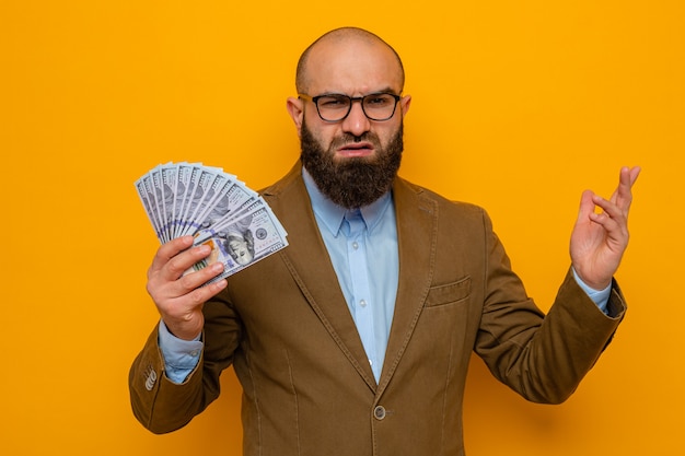 Бородатый мужчина в коричневом костюме в очках держит деньги, глядя в камеру, недовольно поднимая руку, стоя на оранжевом фоне