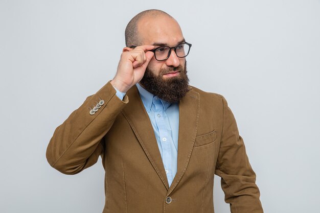 Bearded man in brown suit wearing glasses looking aside with confident expression standing over white background