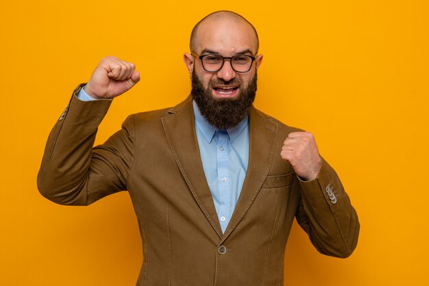 Bearded man in brown suit wearing glasses looking at camera happy and excited clenching fists standing over orange background