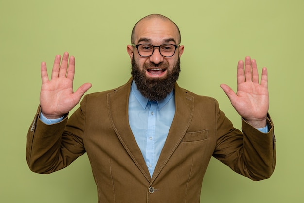Бородатый мужчина в коричневом костюме в очках смотрит в камеру счастливым и взволнованным, поднимая руки, стоя на зеленом фоне