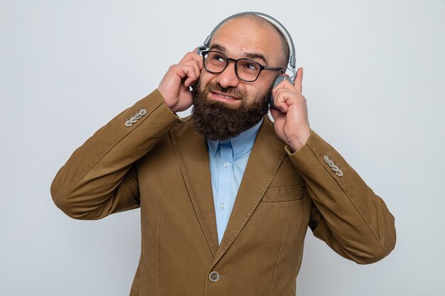 Bearded man in brown suit wearing glasses with headphones looking up smiling enjoying his favorite music standing over white background