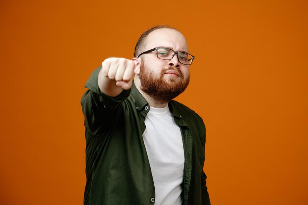 Бородатый мужчина в повседневной одежде в очках сжимает кулак, указывая им на какмеру, выглядит уверенно, стоя на оранжевом фоне