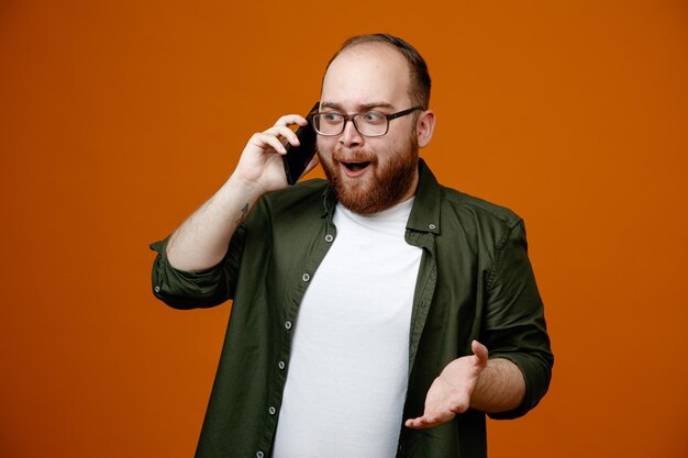 Бородатый мужчина в повседневной одежде в очках счастлив и удивлен, улыбаясь, разговаривая по мобильному телефону, стоя на оранжевом фоне