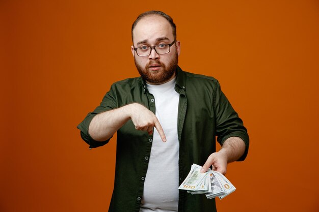 オレンジ色の背景の上に自信を持って立っているお金を人差し指で指している現金を保持している眼鏡をかけたカジュアルな服を着たひげを生やした男