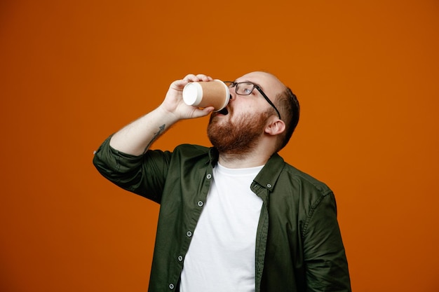 Бородатый мужчина в повседневной одежде в очках держит чашку кофе и пьет кофе, стоя на оранжевом фоне