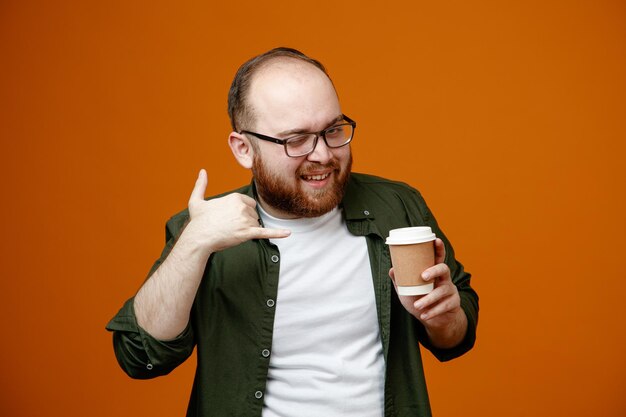 Bearded man in casual clothes wearing glasses holding cup of coffee looking at camera winking and smiling showing call me gesture standing over orange background