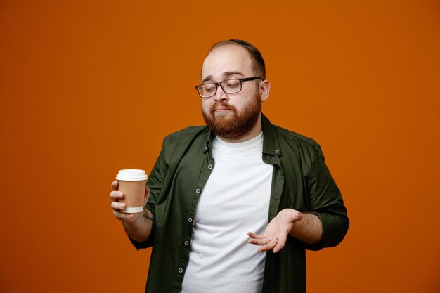 Бородатый мужчина в повседневной одежде в очках держит чашку кофе и смотрит на нее, смущенный скептическим выражением лица, стоящим на оранжевом фоне