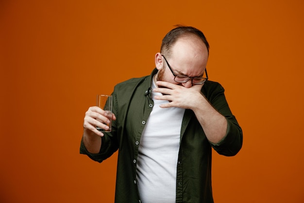 Бородатый мужчина в повседневной одежде в очках, держащий стакан воды, выглядит смущенным и разочарованным, стоя на оранжевом фоне