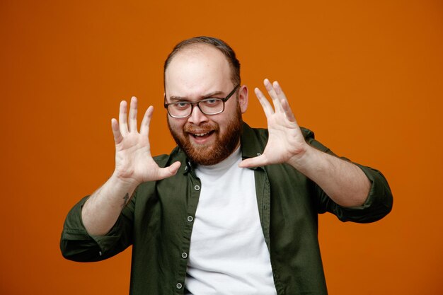 Бородатый мужчина в повседневной одежде в очках смотрит в камеру, пугающе поднимая руки, стоя на оранжевом фоне