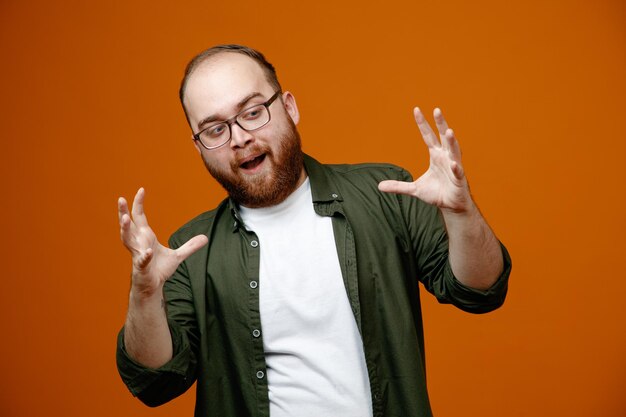 Bearded man in casual clothes wearing glasses looking at camera frightening raising arms standing over orange background