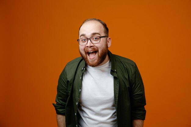 Bearded man in casual clothes wearing glasses looking at camera happy and excited screaming having fun standing over orange background