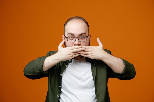 Bearded man in casual clothes wearing glasses looking at camera with serious face covering mouth with hands standing over orange background