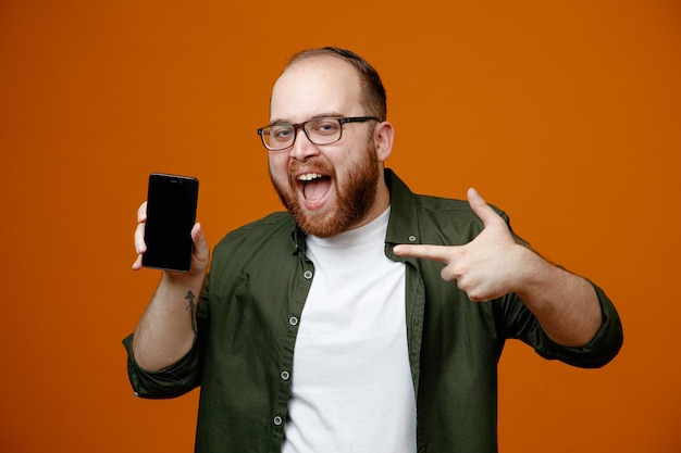 Бородатый мужчина в повседневной одежде в очках показывает смартфон, указывая на него указательным пальцем, весело улыбаясь, стоя на оранжевом фоне