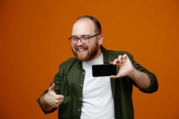 Uomo barbuto in abiti casual con occhiali che mostrano smartphone sorridente allegramente mostrando pollice in piedi su sfondo arancione