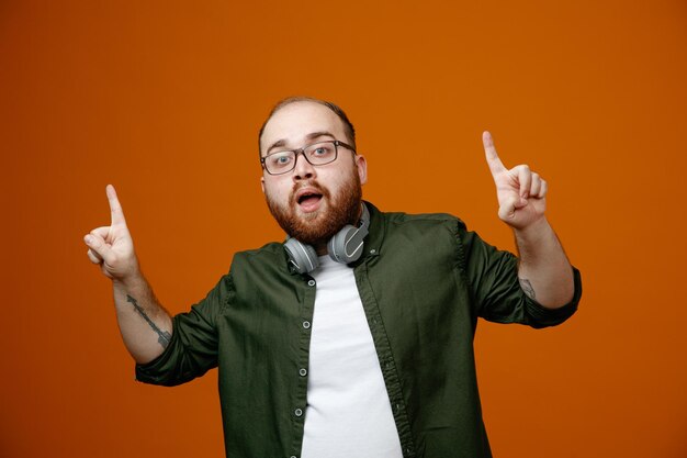 Bearded man in casual clothes wearing glasses with headphones around neck looking at camera happy and surprised pointing with index fingers up standing over orange background
