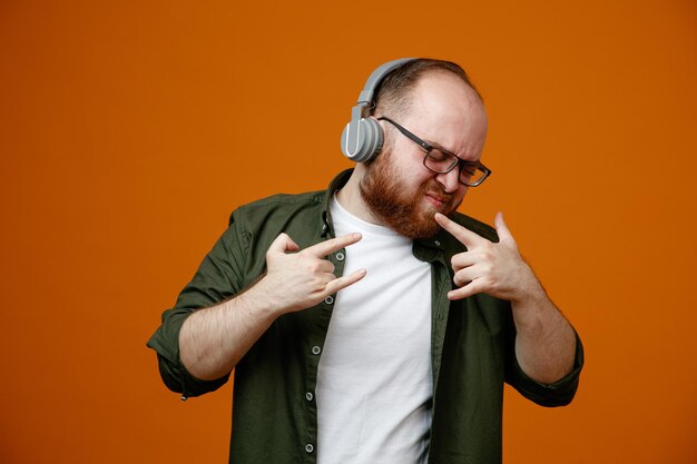 Бородатый мужчина в повседневной одежде в очках с наушниками счастлив и взволнован, показывая рок-символы, слушая рок-музыку, стоящую на оранжевом фоне