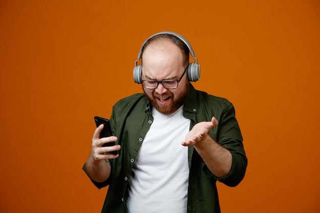 Бородатый мужчина в повседневной одежде в очках с наушниками, держащий смартфон, выглядит сбитым с толку, недовольно поднимая руку, стоя на оранжевом фоне