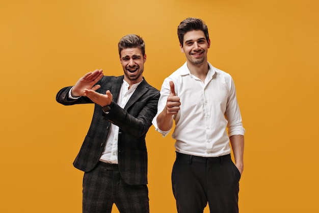 Free photo bearded man in checkered suit claps hands and smiles brunet guy in white shirt shows thumbs up businessmen pose on orange background