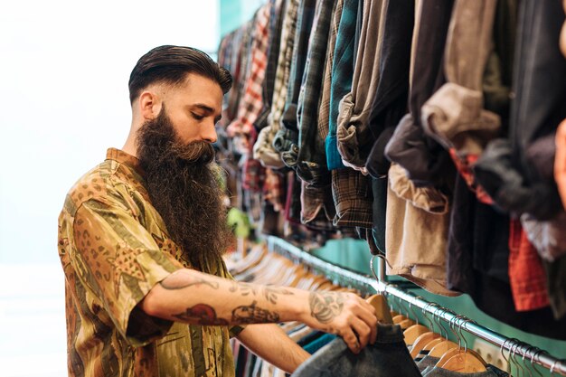 Бородатый мужчина выбирает рубашку висит на рельсе в магазине